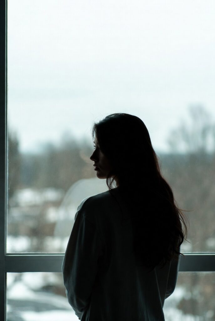 Mulher olhando pela janela em dia nublado representando o intervalo entre identidades durante o cansaço existencial